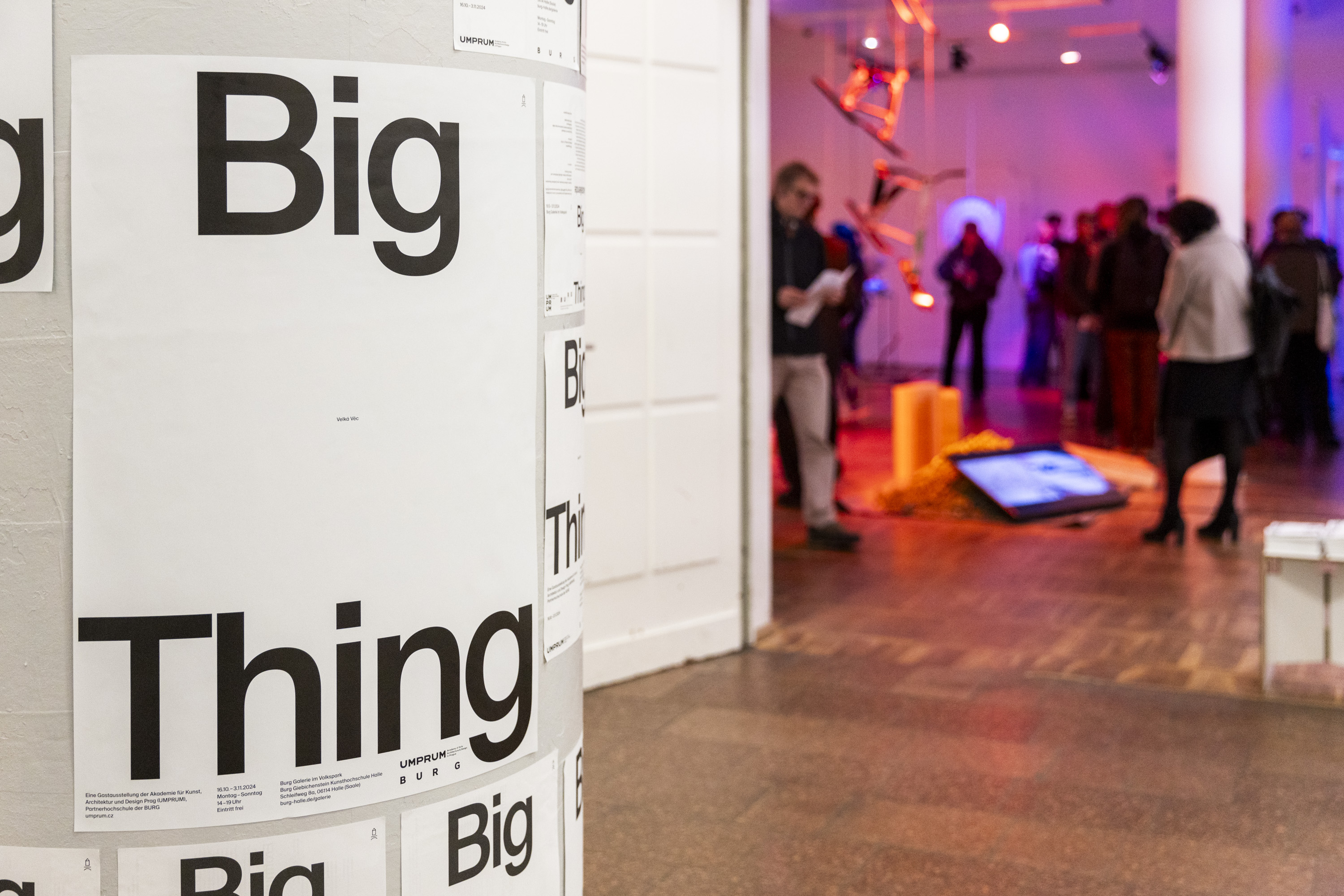 Fotografie einer links im Bild stehenden weißen Litfaßsäule mit einem Plakat zu"Big Thing". Rechtds im Bild sieht man Besucher:innen einer Ausstellung.  