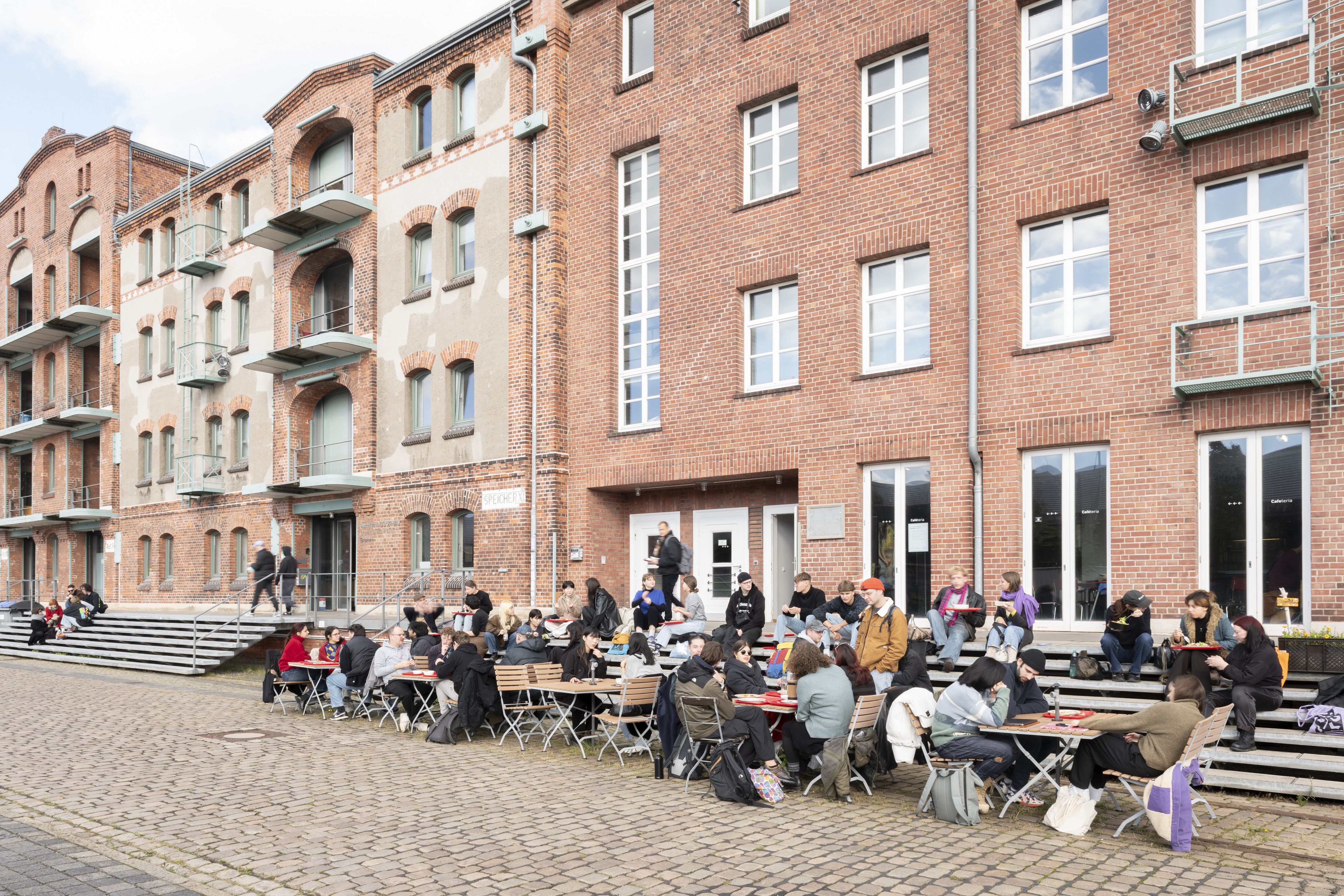 Backsteingebäude der HfK Bremen, vor denen Studierende an Tischen und auf Bänken sitzen. 