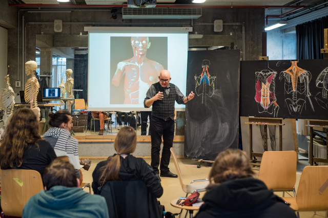 Zu sehen ist eine Unterrichtsszene, in der ein Mann in der Mitte des Raumes steht und den vor ihm sitzenden Studierenden etwas erklärt. Zu sehen sind außerdem Bilder und Zeichnungen der menschlichen Anatomie.