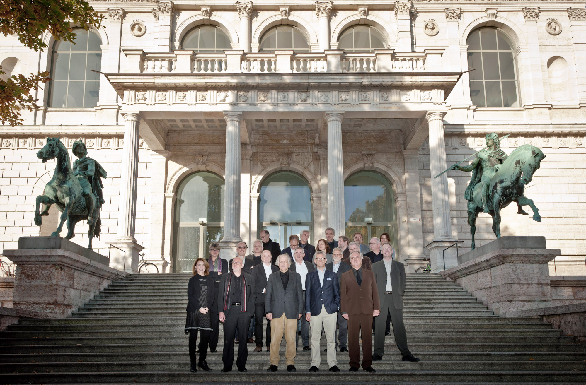 Zu sehen ist ein Gruppenfoto der KHK auf der Treppe vor dem Altbau der AdBK München. Links und rechts der Treppe sind zwei Reiterstatuen zu sehen